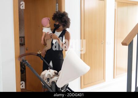 Donna Afro-americana in maschera protettiva in piedi con adorabile bambino vicino carrozza bambino mentre si prepara per passeggiare Foto Stock