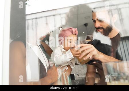 Angolo basso attraverso finestra di coppia multirazziale che dà succo per un bambino carino mentre si siede insieme al tavolo nel caffè Foto Stock