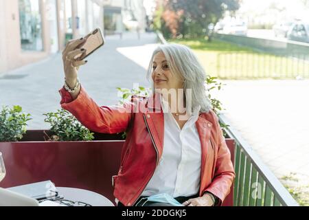 Donna di mezza età felicissima con capelli grigi che prendono selfie su smartphone mentre si siede in una caffetteria all'aperto e si aspetta l'ordine Foto Stock