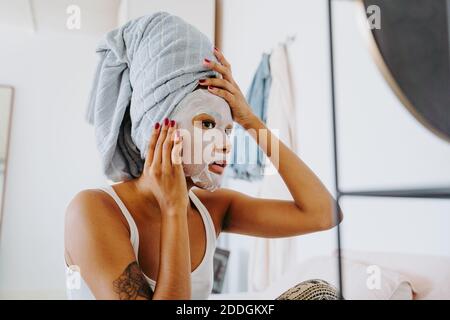 Giovane femmina a specchio con maschera di tessuto idratante per la pelle faccia durante la procedura domestica di bellezza Foto Stock