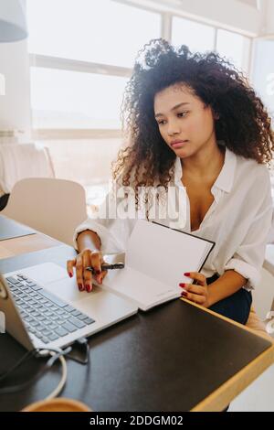 Moderna giovane afroamericana dipendente remoto in abiti casual seduto al tavolo con il computer portatile e scrivendo nel pianificatore mentre lavorare in ufficio domestico Foto Stock