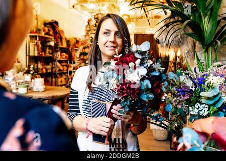 Fiorista femminile professionista che tiene bouquet di fiori mentre aiuta tagliato irriconoscibile cliente in negozio di arredamento guardando l'un l'altro Foto Stock