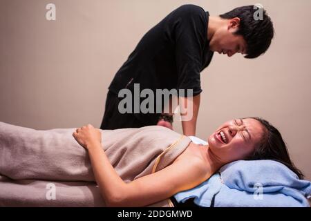 Vista laterale della giovane paziente asiatica che piange nel dolore mentre si ottiene un massaggio osteopatico professionale dal terapeuta in clinica Foto Stock