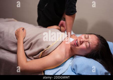 Vista laterale della giovane paziente asiatica che piange nel dolore mentre si ottiene un massaggio osteopatico professionale dal terapeuta in clinica Foto Stock