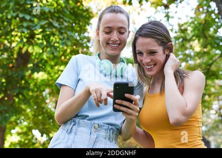 Da sotto allegra giovane ragazza multirazziale utilizzando smartphone e discutere immagini divertenti mentre passeggiando insieme sulla strada urbana in estate da Foto Stock