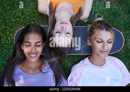 Vista dall'alto di un gruppo di studentesse teenager multirazziali che giace su erba verde con teste sullo skateboard mentre si raffredda insieme nel parco estivo Foto Stock