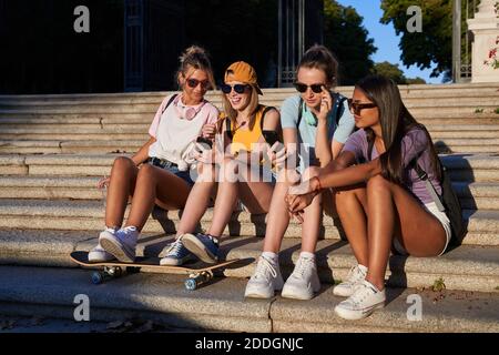 Allegro giovane trendy girlfriends multirazziale in occhiali da sole che naviga telefoni cellulari mentre riposano insieme su scale di pietra nella soleggiata giornata estiva in città Foto Stock