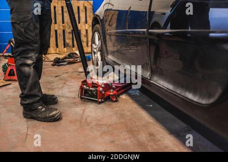 Tecnico irriconoscibile che solleva un veicolo moderno con martinetto idraulico durante il lavoro in garage professionale Foto Stock