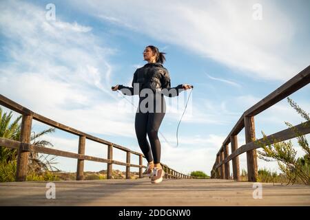 Da sotto il corpo completo di fit femminile sportivo in nero outfit sportivo che salta con la corda saltante su passerella in legno durante allenamento fitness all'aperto in sintesi Foto Stock