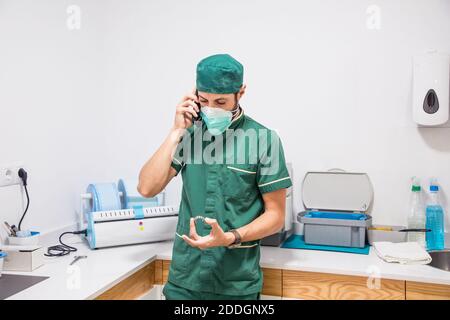 Dentista maschile in maschera e uniforme parlando su smartphone mentre tenere un getto dentale in un moderno ospedale dentale Foto Stock