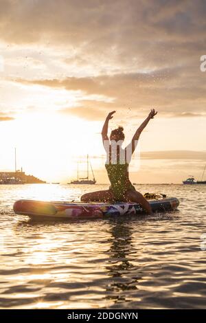 Allegro slim surfer femminile seduta su tavola galleggiante e. spruzzi d'acqua contro il cielo del tramonto mentre godendo le vacanze estive a. mare Foto Stock