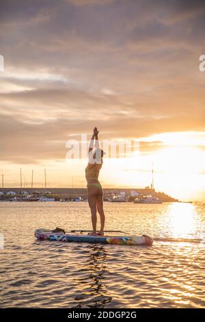Profilo con vista laterale di una femmina sottile in posizione di montagna con le braccia in su mentre si pratica yoga su tavola galleggiante contro il cielo del tramonto Foto Stock