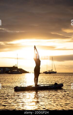 Profilo con vista laterale di una femmina sottile in posizione di montagna con le braccia in su mentre si pratica yoga su tavola galleggiante contro il cielo del tramonto Foto Stock