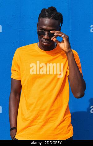 Serio giovane afroamericano ragazzo in maglietta gialla luminosa e occhiali da sole alla moda che guardano lontano su sfondo blu Foto Stock
