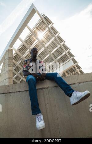 Da sotto il corpo pieno di elegante ragazzo nero in trendy camicia colorata e jeans con sneaker bianche in cemento recinzione contro l'edificio moderno della città Foto Stock