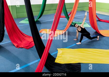 Alto angolo di uomo flessibile in leotard facendo esercizio stretching vicino a colorate sete aeree durante le prove in studio Foto Stock