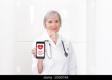 Medico senior amichevole con uno smartphone che mostra un'app di agenda della pressione sanguigna, sfondo bianco, spazio di copia. Foto Stock