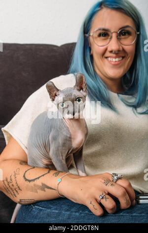Donna allegra con capelli tinti e gatto Sphynx guardando la macchina fotografica mentre riposa sul divano a casa Foto Stock