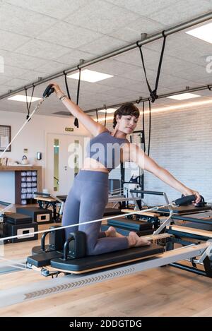Fit adulta femmina facendo inginocchiare Lean Back esercizio con outtensed Armi sul riformatore durante l'allenamento Pilates in palestra Foto Stock