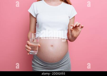 Donna incinta che tiene un bicchiere d'acqua e pillole, vitamine per la gravidanza, la futura madre si aspetta un bambino, su uno sfondo rosa Foto Stock