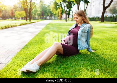 Felice giovane donna incinta, si siede nel parco sul prato, guarda il ventre e lo abbraccia con le mani Foto Stock