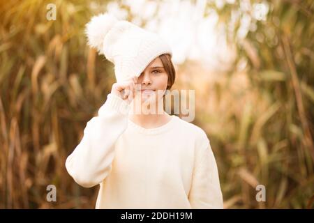 Divertente ragazza teen sorride tirando il cappuccio sopra gli occhi. Ritratto di divertente ragazza carina nascosto i suoi occhi in bianco inverno lavorato a maglia cappello e gioioso sorridente Foto Stock