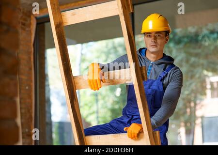 Giovane costruttore maschile in tute e elmetto guardando la macchina fotografica, salendo la scala mentre lavorando al cantiere. Costruzione, persone, concetto di professione Foto Stock