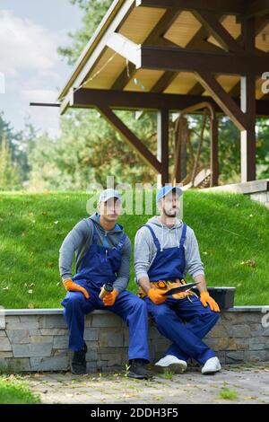 Scatto a tutta lunghezza di due giovani operai in uniforme che prendono una pausa, seduti all'aperto con caffè mentre lavorano al progetto di costruzione. Costruzione, partnership, concetto di professione Foto Stock