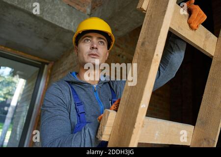 Ritratto di attraente giovane costruttore maschile in elmetto che guarda via, salendo la scala mentre lavora in cantiere. Costruzione, persone, concetto di professione Foto Stock