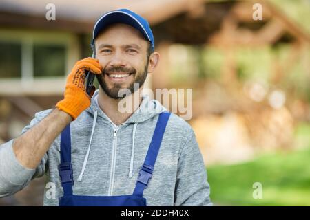 Ritratto di gioioso giovane costruttore maschile in uniforme sorridente mentre si parla al telefono, in piedi all'aperto in una calda giornata di sole. Professione, comunicazione e concetto di costruzione Foto Stock