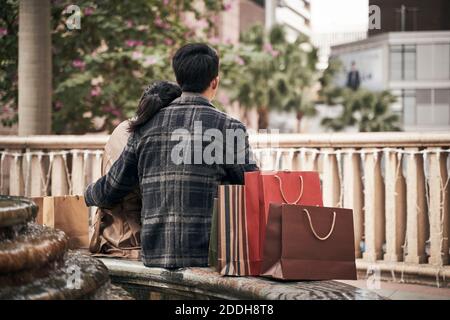 vista posteriore della giovane coppia asiatica riposante all'aperto mentre shopping in città Foto Stock