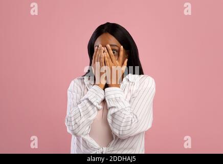 Ritratto di giovane donna nera che ricopre il viso con le mani, sentendosi impaurito o stressato sullo sfondo rosa dello studio Foto Stock