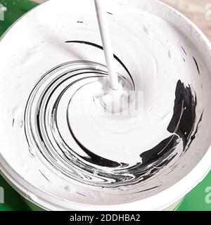 Il pittore della casa sta mescolando la vernice nera e bianca nel secchio usando il miscelatore della pittura per ottenere un colore grigio. Foto Stock
