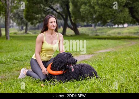 La barba nera è adagiata sull'erba con l'anello del giocattolo sul collo e la proprietaria femminile vicino al parco pubblico. Cane con capelli lunghi si trova all'aperto. Foto Stock