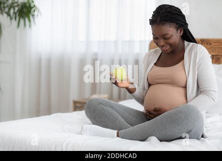 Nutrizione delle donne incinte. Donna incinta nera seduta sul letto, tenendo mela verde Foto Stock