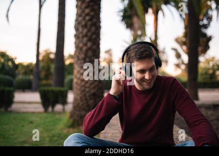 Biondo ragazzo che ascolta la musica con una cuffia wireless seduta sul pavimento. Ascoltare la canzone. Concetto musicale. Pullover rosso. Foto Stock