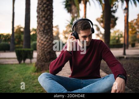 Biondo ragazzo che ascolta la musica con una cuffia wireless seduta sul pavimento. Ascoltare la canzone. Concetto musicale. Pullover rosso. Foto Stock