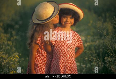 due sorelle ascoltate in cappelli di paglia in piedi in un prato a margherita al tramonto Foto Stock