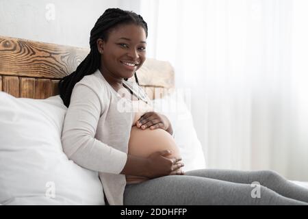 Donna incinta nera sorridente riposante sul letto e tenendo le mani Su Belly Foto Stock