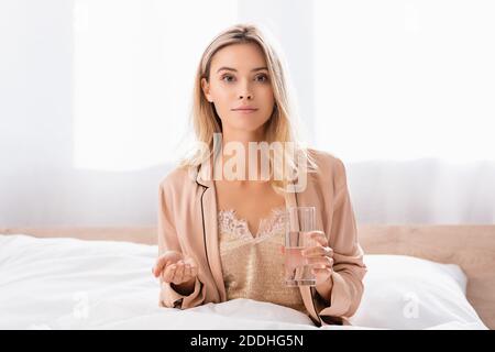 Donna in pigiama guardando la macchina fotografica mentre tiene il vetro di acqua e puntando con la mano sul letto Foto Stock