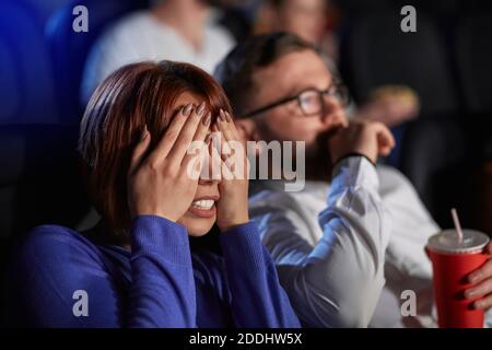 Fuoco selettivo di giovane ragazza spaventata che copre gli occhi con le mani, guardando film di horror nel cinema. Vista laterale dell'uomo caucasico spaventato e della donna rossa con testa che gode di un film spaventoso. Foto Stock