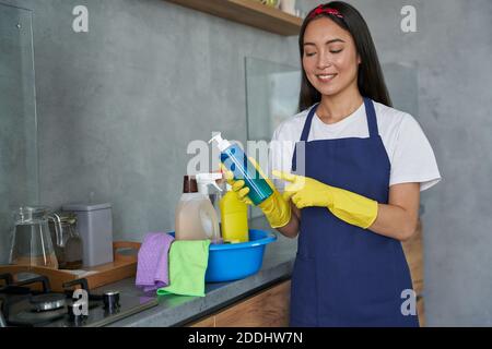 Giovane donna gioiosa, donna che si pulisce indossando guanti protettivi, sorridendo e tenendo in mano il prodotto per la pulizia della casa mentre si prepara per la pulizia della casa. Concetto di servizio di pulizia e pulizia Foto Stock