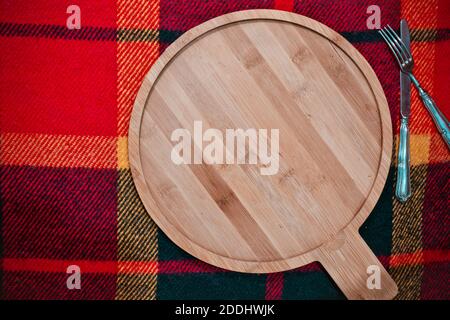 Fondo del menu della stagione fredda, cottura o cottura: Tagliere di legno rotondo vuoto su panno a scacchi con coltello e forchetta Foto Stock