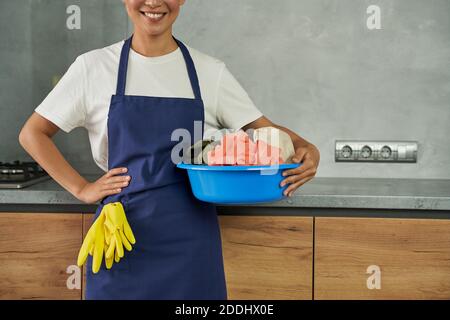 Pronto per lavori domestici. Un piccolo colpo di giovane donna allegra, una donna che pulisce sorridendo alla macchina fotografica mentre tiene il cesto di plastica con la lavanderia sporca all'interno. Concetto di servizio di pulizia professionale della casa Foto Stock