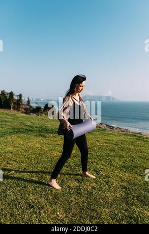 Donna si prepara a mettere il suo tappeto yoga sull'erba di fronte al mare. Spazio di copia Foto Stock