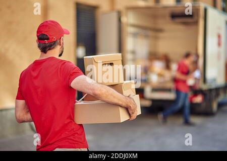 Vista posteriore di un corriere maschile in uniforme rossa che tiene pacchi per la consegna in piedi presso il negozio, fuoco selettivo. Concetto di servizi di consegna Foto Stock