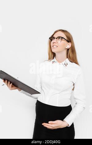 Immagine di una giovane donna incinta di affari con gli occhiali, la ragazza pensiva tiene una cartella con i documenti in mano e scrive i rapporti, isolato su backgr bianco Foto Stock