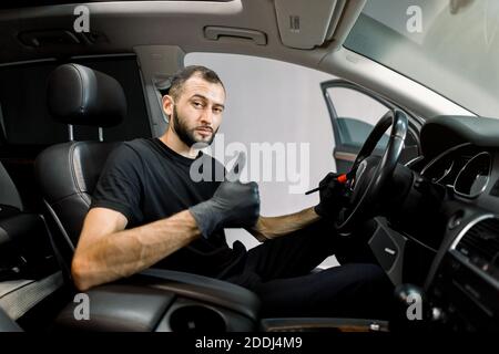 Giovane uomo caucasico autolavaggio dettaglio lavoratore utilizzando la spazzola di pulizia e rimuovere la polvere dal pannello di controllo auto e volante, mostrando il pollice verso l'alto Foto Stock