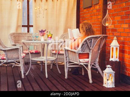 Balcone con vista sul mare. Donna che legge un libro di carta sul balcone della casa all'aperto. Luci a corda che illuminano lo spazio. Molte candele e lanterne. Foto Stock