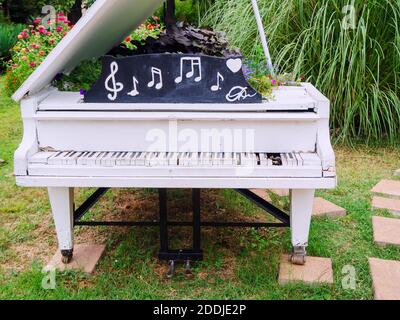 Grande pianoforte decorativo bianco con tasti rotti e note dipinte in cima e decorata con fiori si erge nel parco con fiori e piante Foto Stock